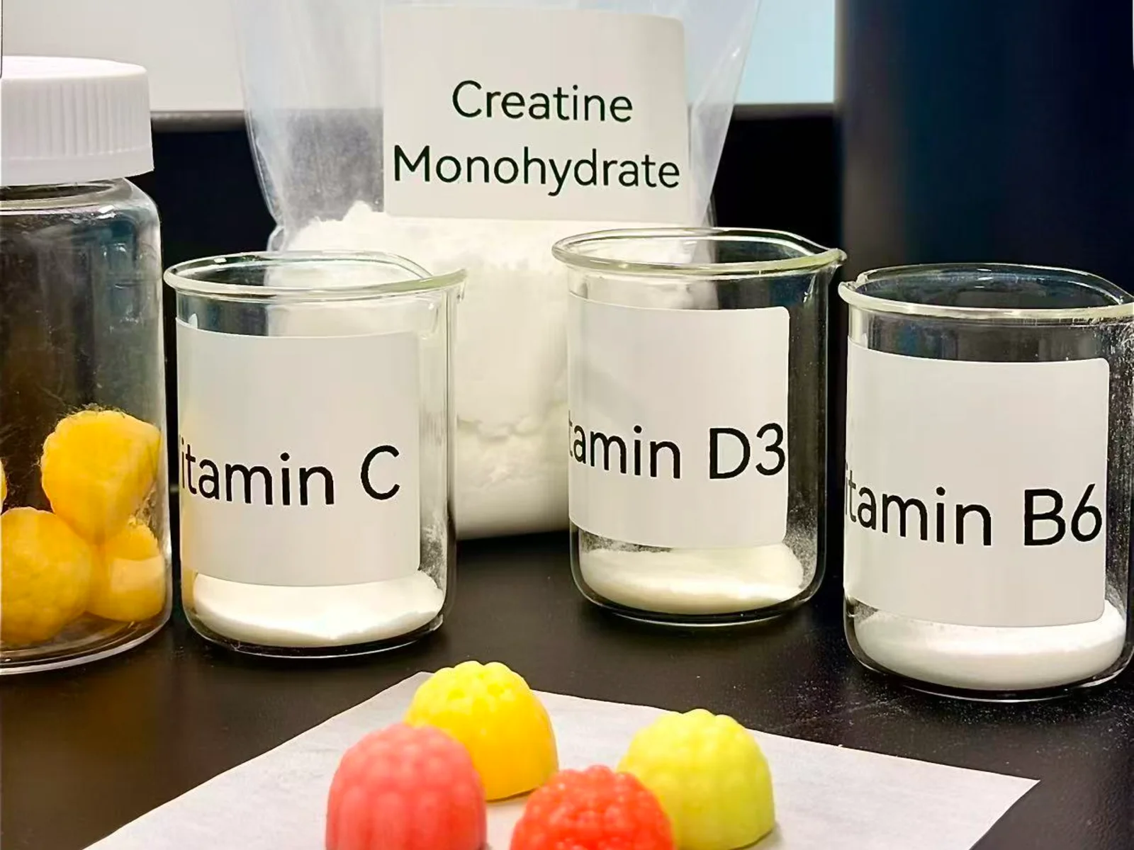 Gummy samples beside a bag labeled creatine monohydrate and beakers labeled vitamins C, D3, and B6, illustrating ingredient options for gummy formulation development.