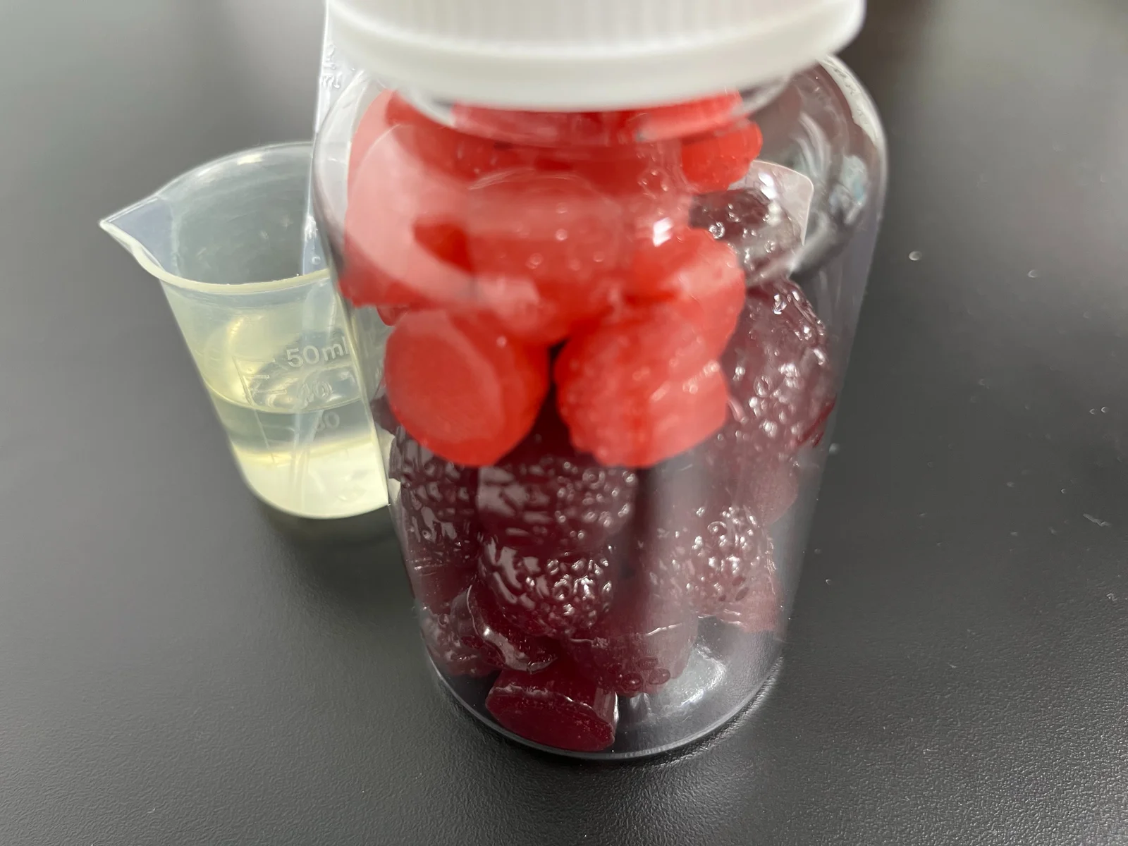Red gummy samples inside a clear bottle with a white cap beside a laboratory beaker, illustrating bottled magnesium gummy packaging and everyday convenience.