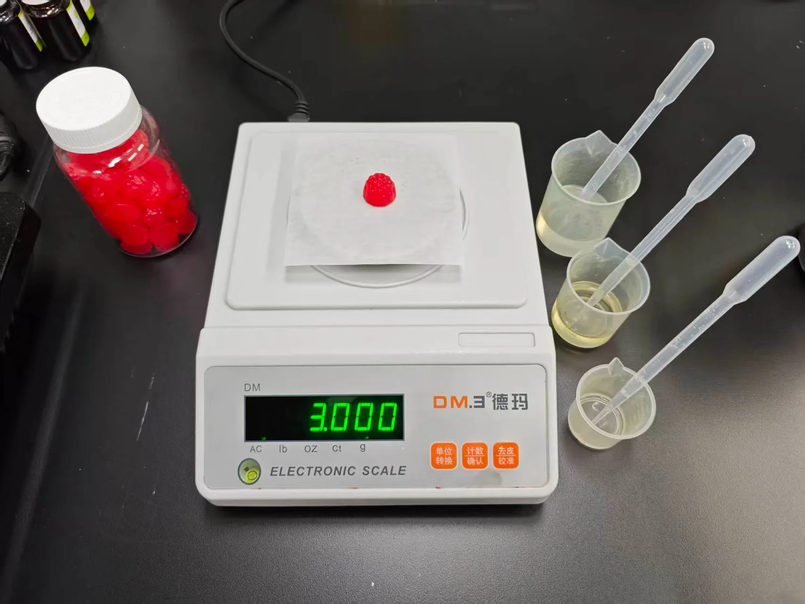 A red gummy on a digital scale beside beakers and transfer pipettes, illustrating sample weight checks when defining magnesium gummy product specifications.